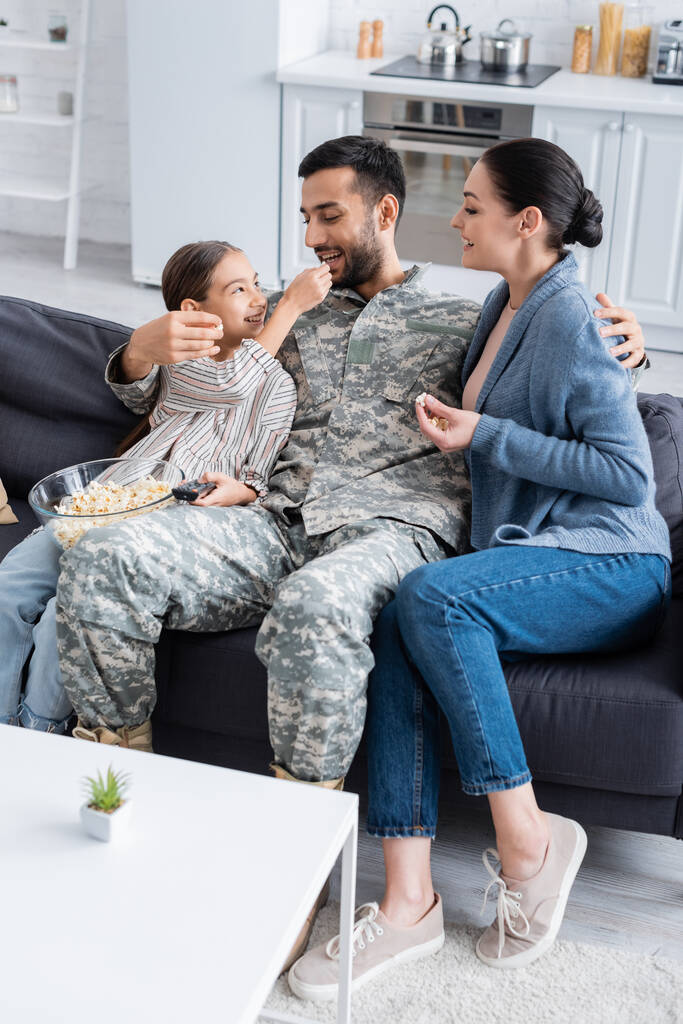 Малюк тримає попкорн і пульт дистанційного керування біля батька у військовій формі і мама на дивані
  - Фото, зображення