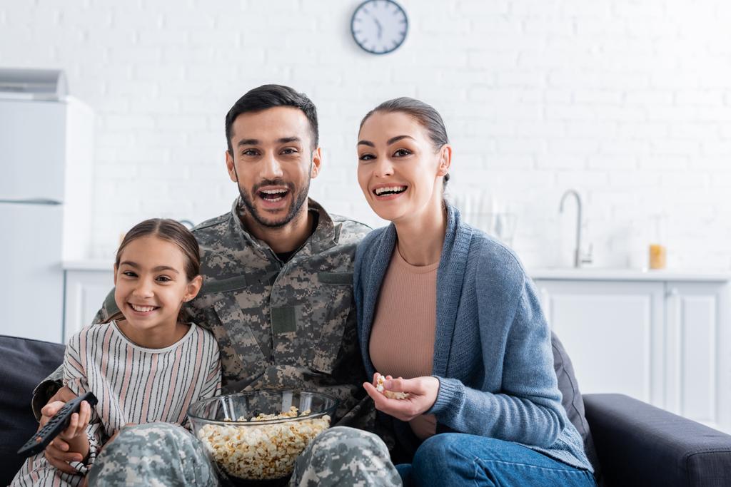 Hombre positivo en uniforme militar sentado cerca de la familia con mando a distancia y palomitas de maíz en el sofá  - Foto, Imagen
