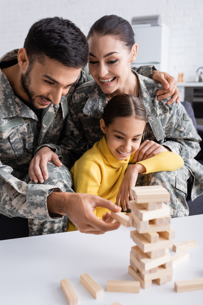 Positive parents in camouflage playing wood blocks game with kid at home  - Photo, Image