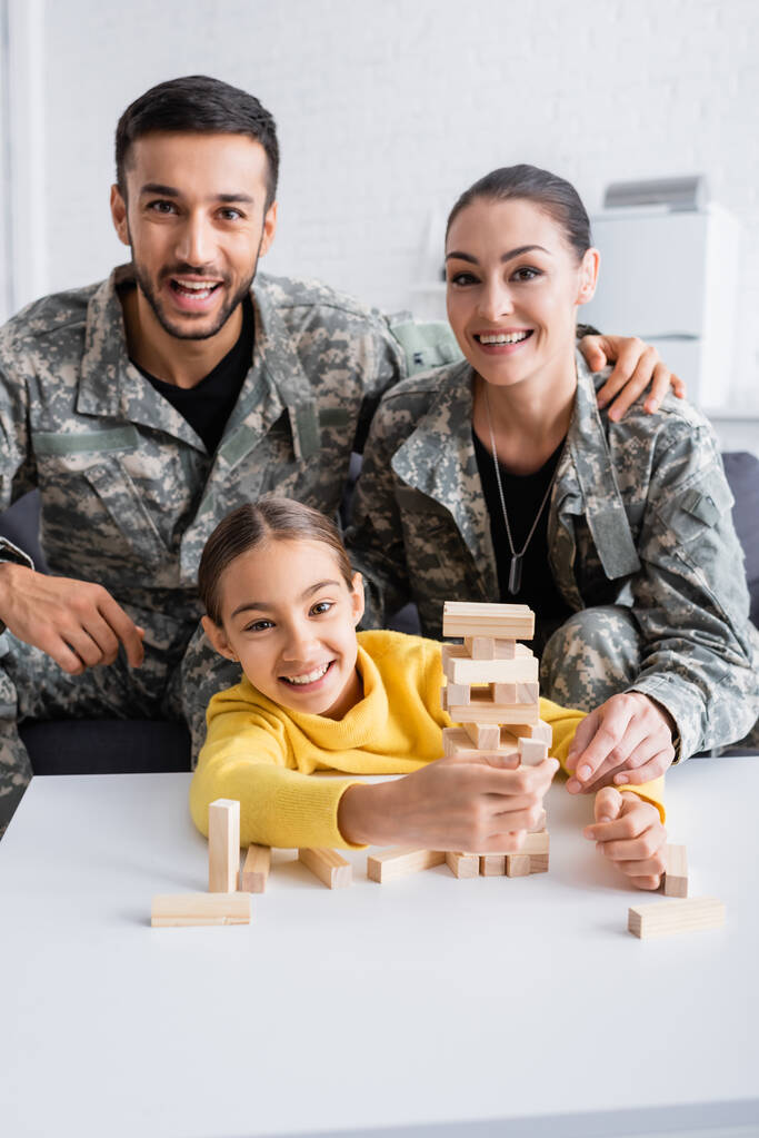 Askeri üniformalı mutlu ebeveynler evlerinde tahta bloklama oyunu oynayan çocuğun yanındaki kameraya bakıyor.  - Fotoğraf, Görsel