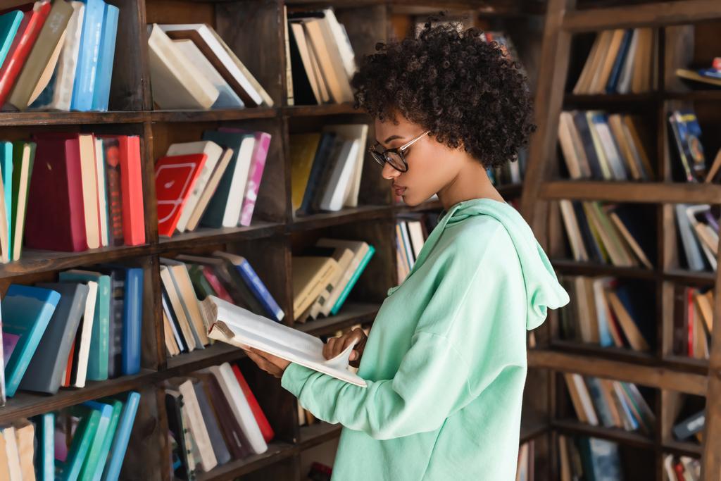 joven estudiante afroamericano en anteojos leyendo libro cerca de librería  - Foto, Imagen