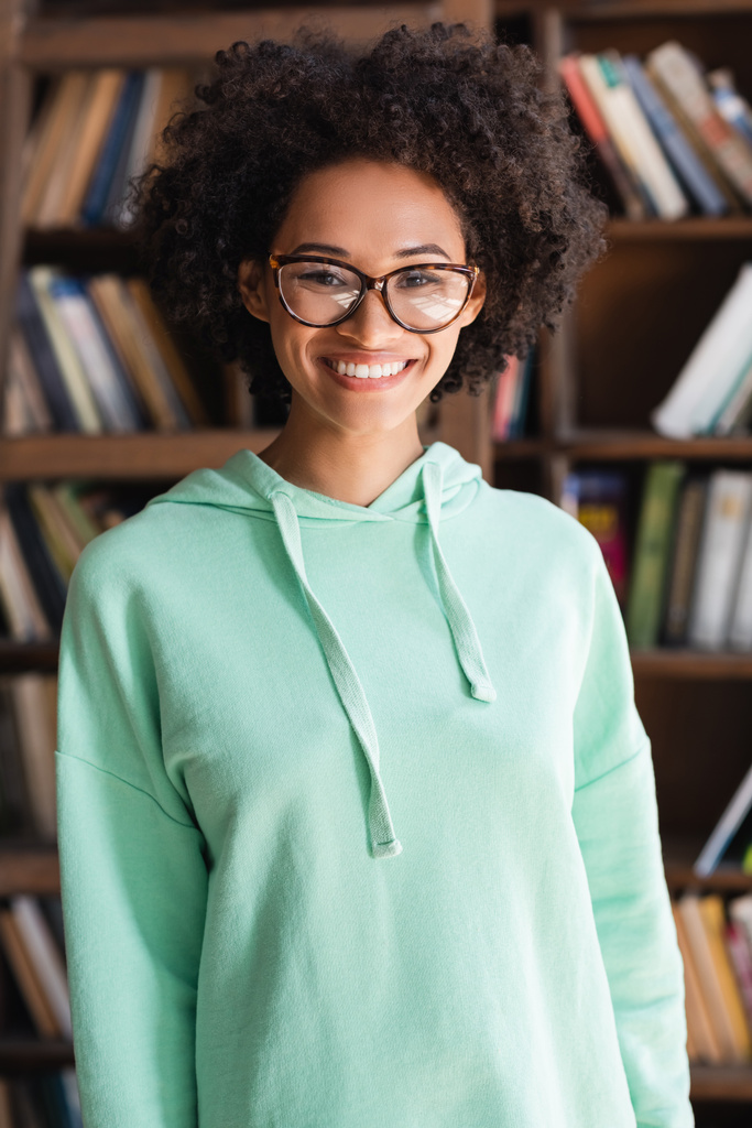 joyeux afro-américain étudiant en lunettes regardant la caméra près de la bibliothèque - Photo, image