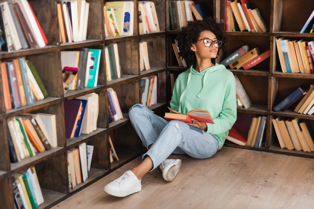 giovane studente afroamericano in occhiali seduto sul pavimento con libro in biblioteca  - Foto, immagini