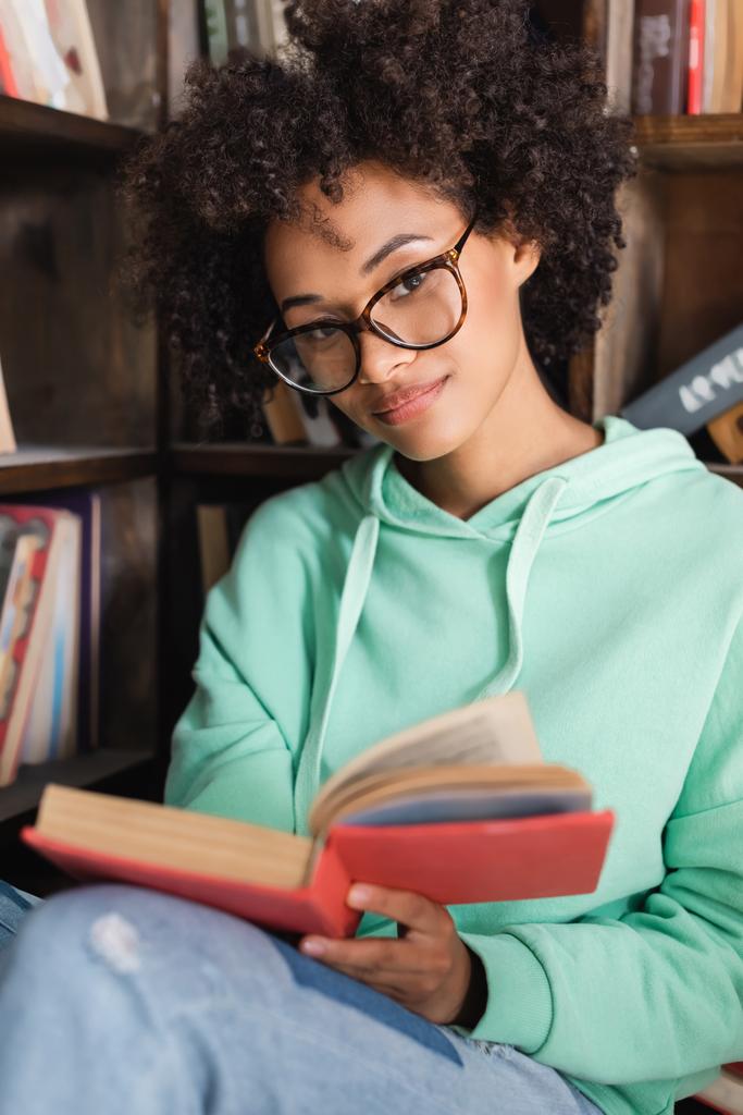 νεαρός Αφροαμερικάνος φοιτητής με γυαλιά ηλίου κρατώντας βιβλίο στη βιβλιοθήκη  - Φωτογραφία, εικόνα