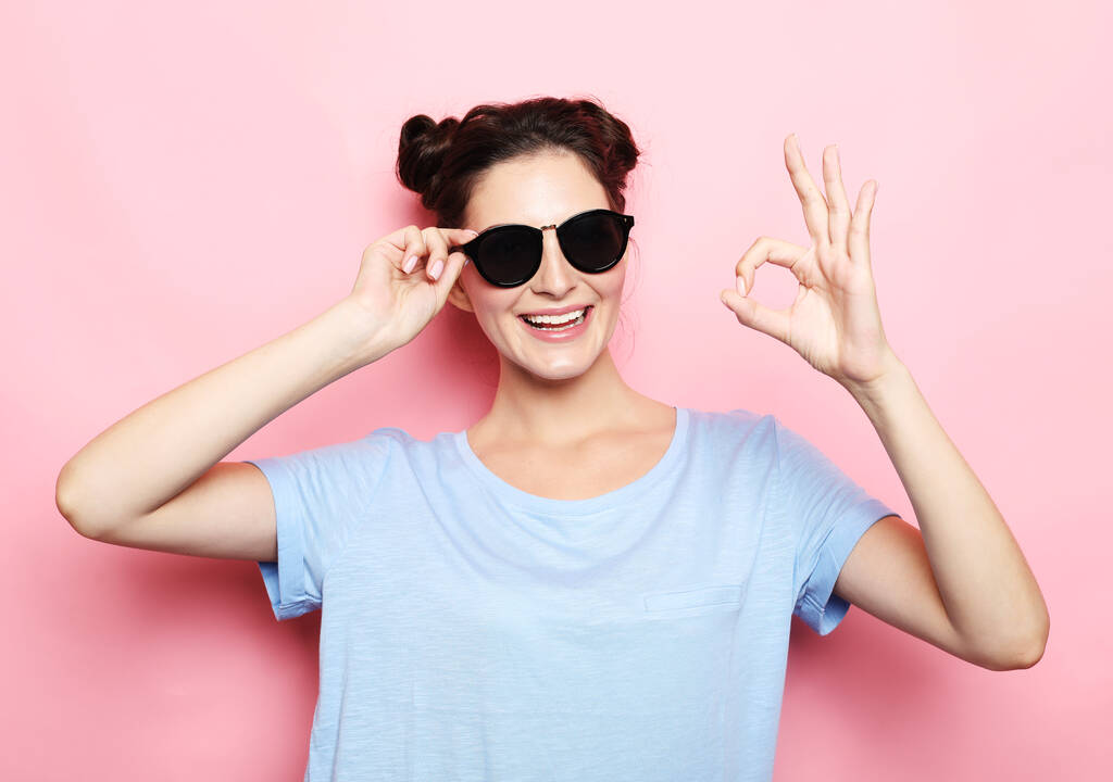 młoda szczęśliwa brunetka kobieta w okularach przeciwsłonecznych noszenie niebieski t-shirt pokazać ok znak na różowym tle - Zdjęcie, obraz