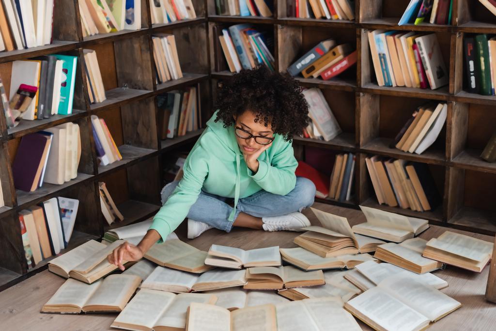 estudiante afroamericano inteligente en anteojos sentado rodeado de libros en la biblioteca  - Foto, imagen