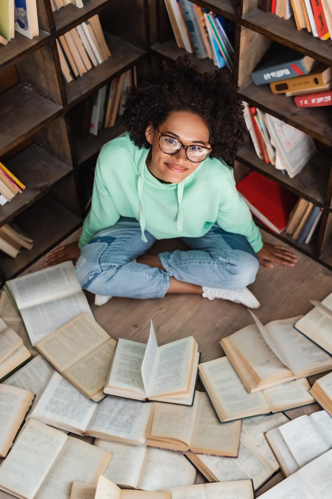 θέα υψηλής γωνίας του ευτυχισμένου Αφροαμερικανού φοιτητή με γυαλιά ηλίου που κάθεται με σταυρωμένα πόδια και περιβάλλεται από βιβλία στη βιβλιοθήκη  - Φωτογραφία, εικόνα