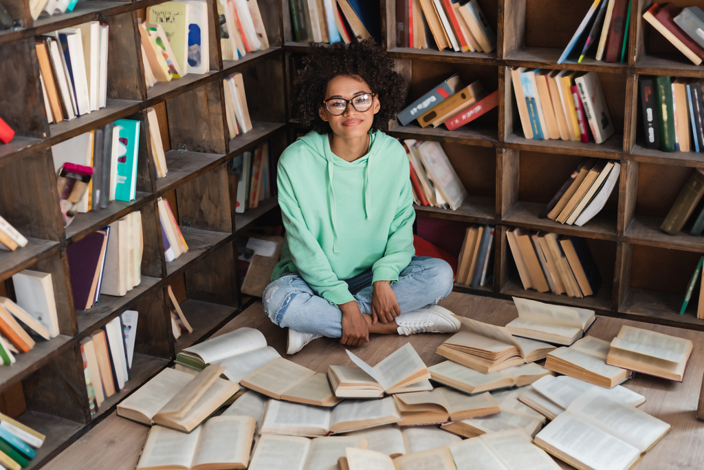 heureux étudiant afro-américain dans des lunettes assis avec les jambes croisées entouré de livres dans la bibliothèque  - Photo, image