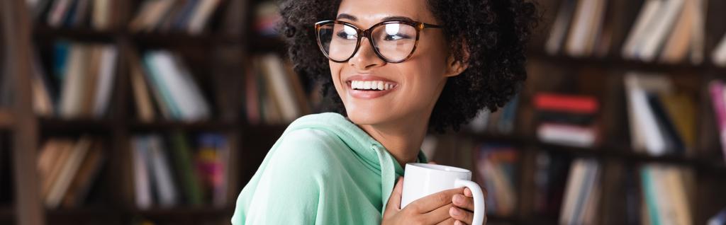 Χαρούμενη νεαρή Αφροαμερικανή με γυαλιά που κρατάει καφέ, πανό. - Φωτογραφία, εικόνα