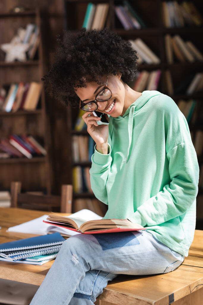 vrolijk Afrikaans amerikaans student in bril praten op mobiele telefoon en lezen boek en zitten op bureau - Foto, afbeelding