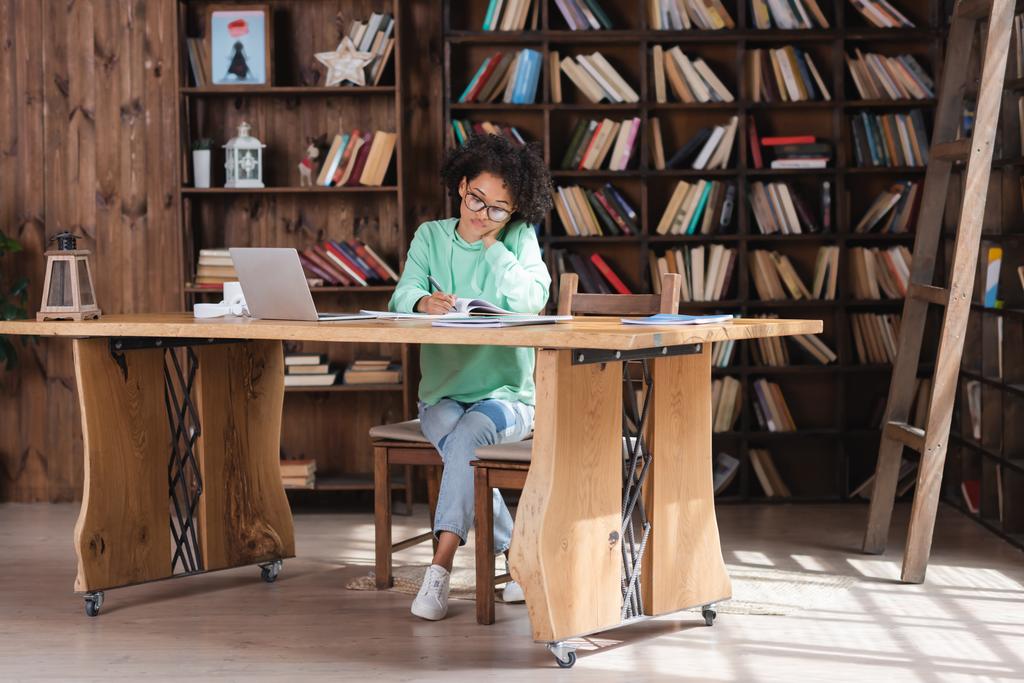 curly african american student in eyeglasses writing in notebook near laptop  - Photo, Image