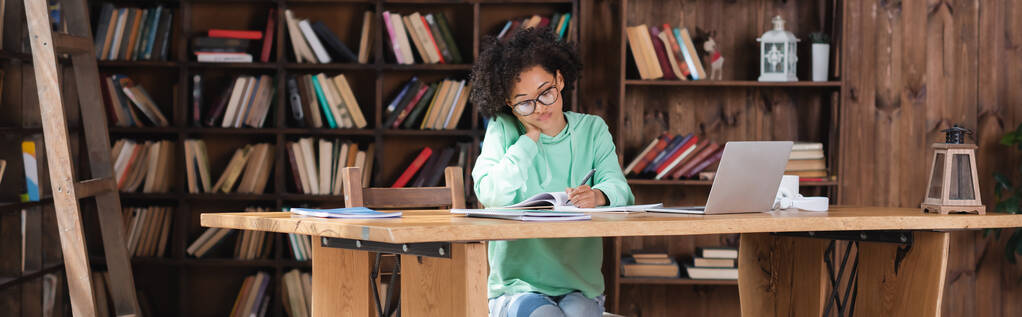 göndör afro-amerikai diák szemüveges írás notebook, miközben tanul online közelében laptop, banner - Fotó, kép