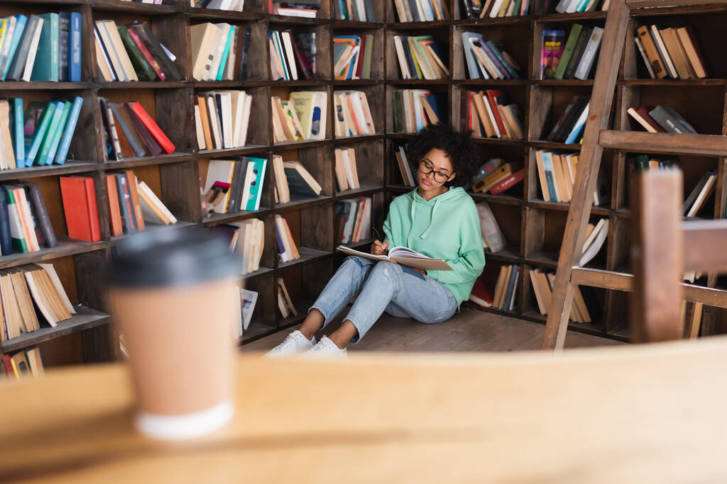 joven estudiante afroamericano en anteojos sentado en el suelo y escribiendo en un cuaderno cerca de la taza de papel borrosa en primer plano - Foto, imagen