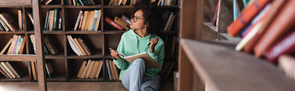σκεπτικός Αφροαμερικανός φοιτητής με γυαλιά ηλίου κάθεται και κρατώντας σημειωματάριο στη βιβλιοθήκη, πανό - Φωτογραφία, εικόνα