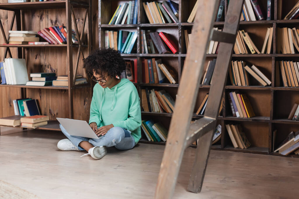 кучерявий афроамериканський студент в окулярах, використовуючи ноутбук, сидячи на підлозі в бібліотеці
 - Фото, зображення