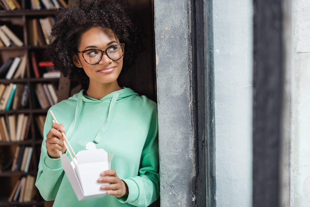 uśmiechnięta Afroamerykanka w okularach trzymająca pałeczki przy pudełku z jedzeniem na wynos  - Zdjęcie, obraz