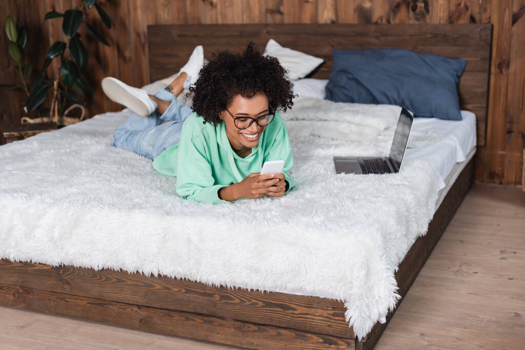 boldog afro-amerikai nő szemüvegben feküdt az ágyban, miközben az okostelefon közelében laptop - Fotó, kép