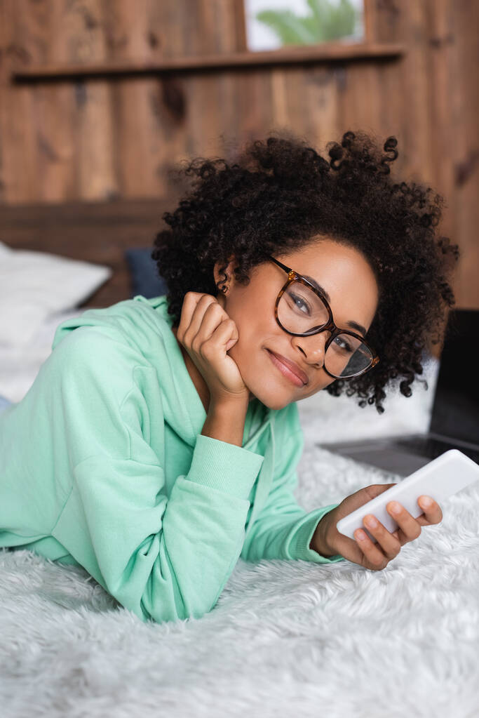 usmívající se africká americká žena v brýlích ležící na posteli, zatímco drží smartphone  - Fotografie, Obrázek