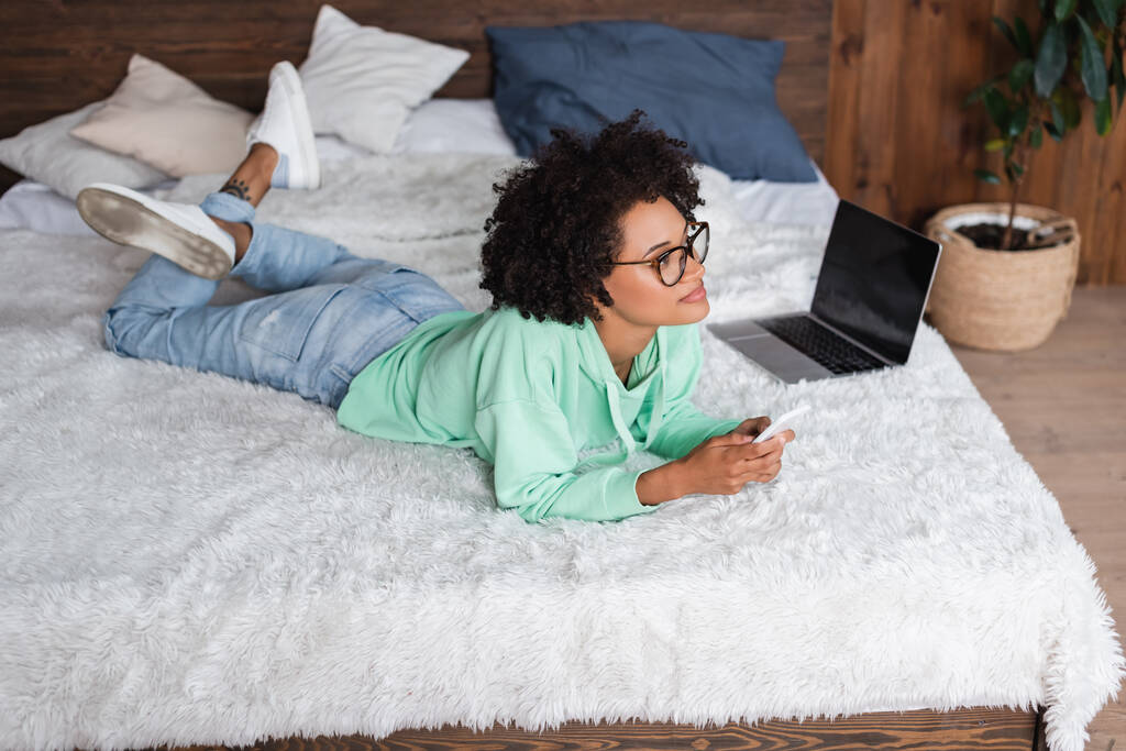 jovem afro-americana em óculos deitado na cama e segurando smartphone perto de laptop - Foto, Imagem