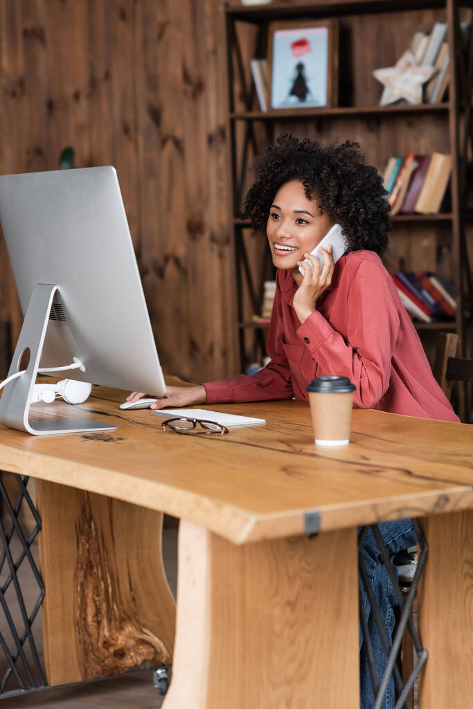 joyeuse femme afro-américaine parlant sur smartphone près du moniteur, tasse en papier et lunettes sur le bureau - Photo, image