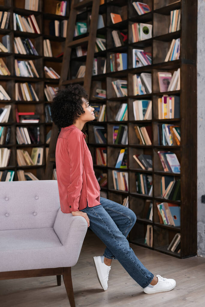 side view of african american woman leaning on grey couch near bookshelf  - Photo, Image