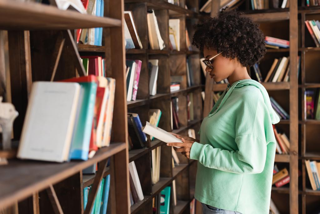 vista laterale dello studente afroamericano in occhiali libro di lettura vicino alla libreria sfocata in primo piano - Foto, immagini