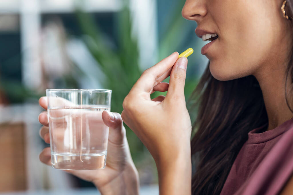 close-up van jonge vrouw het nemen van pillen terwijl het houden van glas water zitten op de bank thuis. - Foto, afbeelding
