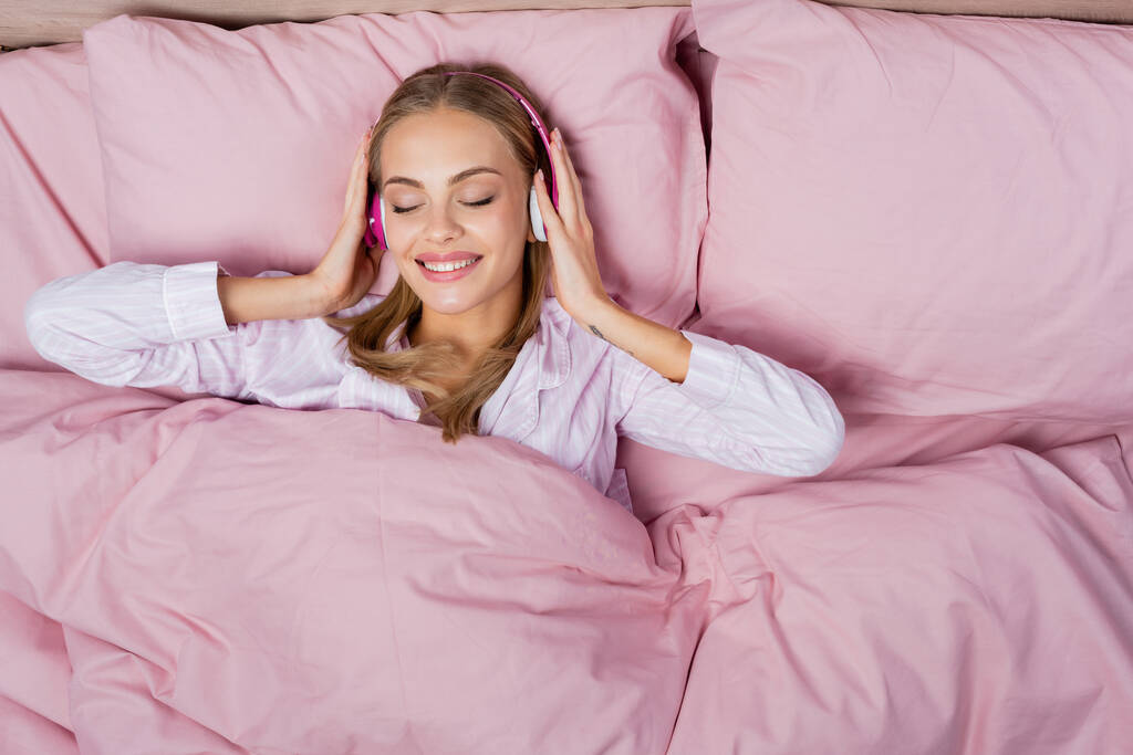 Bovenaanzicht van vrolijke vrouw die muziek luistert in een hoofdtelefoon terwijl ze op bed ligt  - Foto, afbeelding