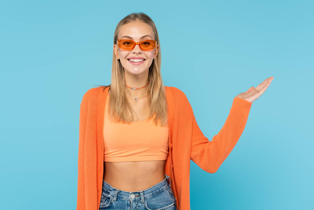 Hymyilevä nainen oranssi aurinkolasit osoittaa käsin eristetty sininen  - Valokuva, kuva