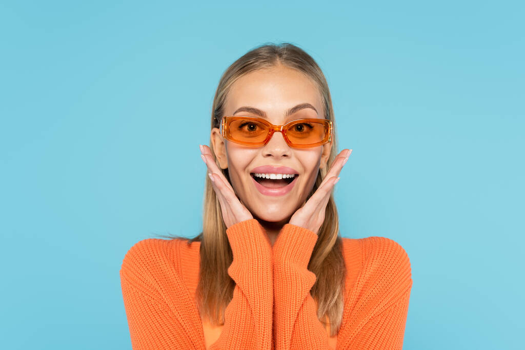 Podekscytowana blondynka w okularach przeciwsłonecznych patrząc na kamerę odizolowaną na niebiesko  - Zdjęcie, obraz