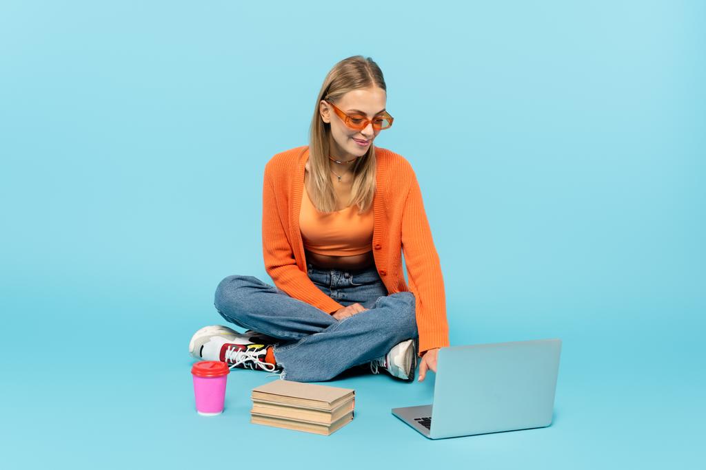 Положительный студент в солнечных очках с помощью ноутбука возле книг и бумажных стаканчиков на синем фоне  - Фото, изображение