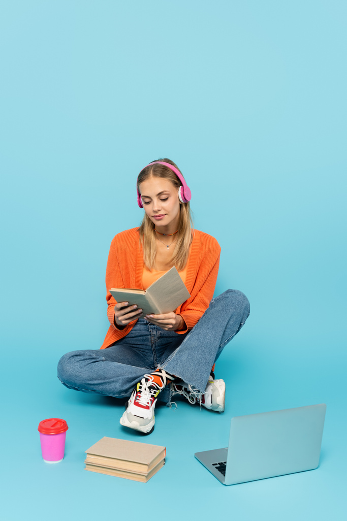 Blondynka w słuchawkach czyta książkę w pobliżu papierowej filiżanki i laptopa na niebieskim tle  - Zdjęcie, obraz