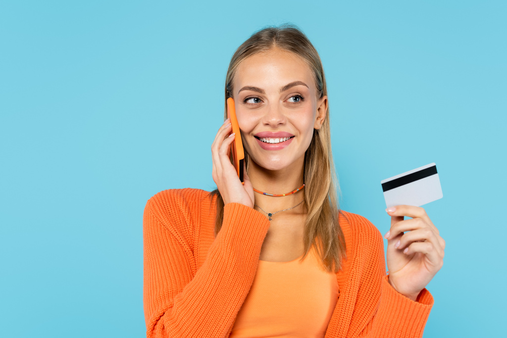 Positiivinen blondi nainen puhuu älypuhelimella ja pitää luottokortin eristetty sininen  - Valokuva, kuva