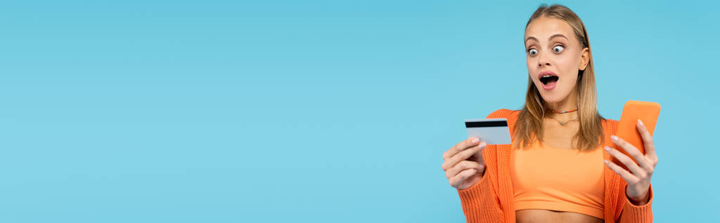 Возбужденная женщина смотрит на кредитную карту, держа мобильный телефон изолирован на синий, баннер  - Фото, изображение
