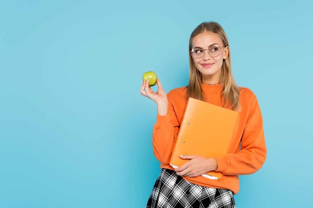 Θετικός μαθητής σε γυαλιά ηλίου που κρατά το φάκελο χαρτιού και το μήλο απομονωμένο στο μπλε  - Φωτογραφία, εικόνα