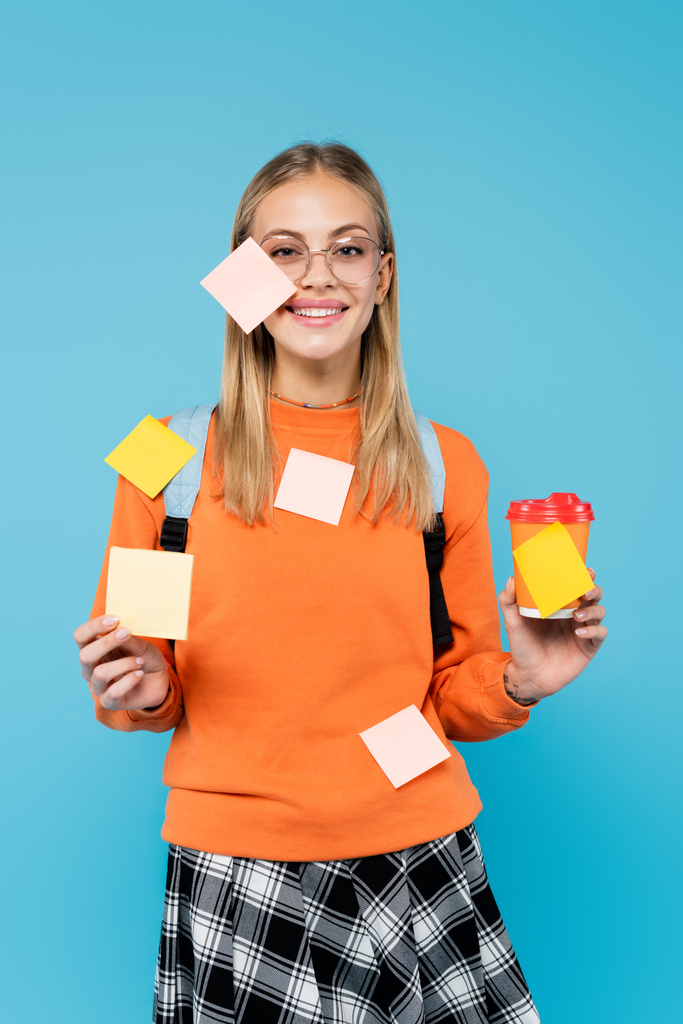 Smiling student in sticky notes holding paper cup isolated on blue  - Photo, Image