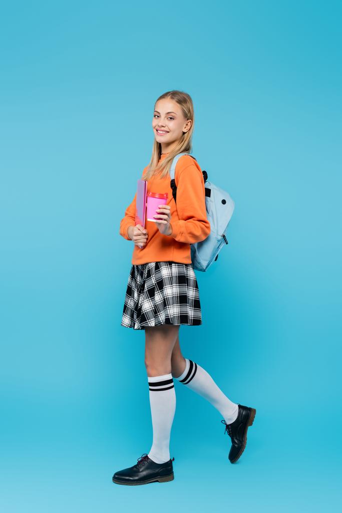 Lächelnder Student mit Rucksack mit Laptop und Kaffee auf blauem Hintergrund  - Foto, Bild