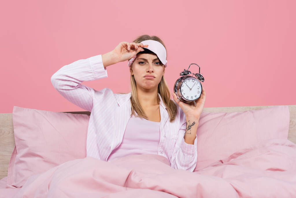 Upset woman holding sleep mask and alarm clock on bed isolated on pink  - Photo, Image