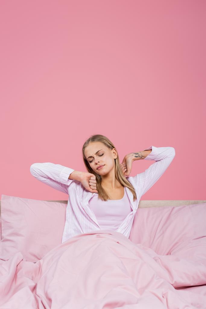 Mladé blondýny žena v pyžamu strečink na posteli izolované na růžové  - Fotografie, Obrázek