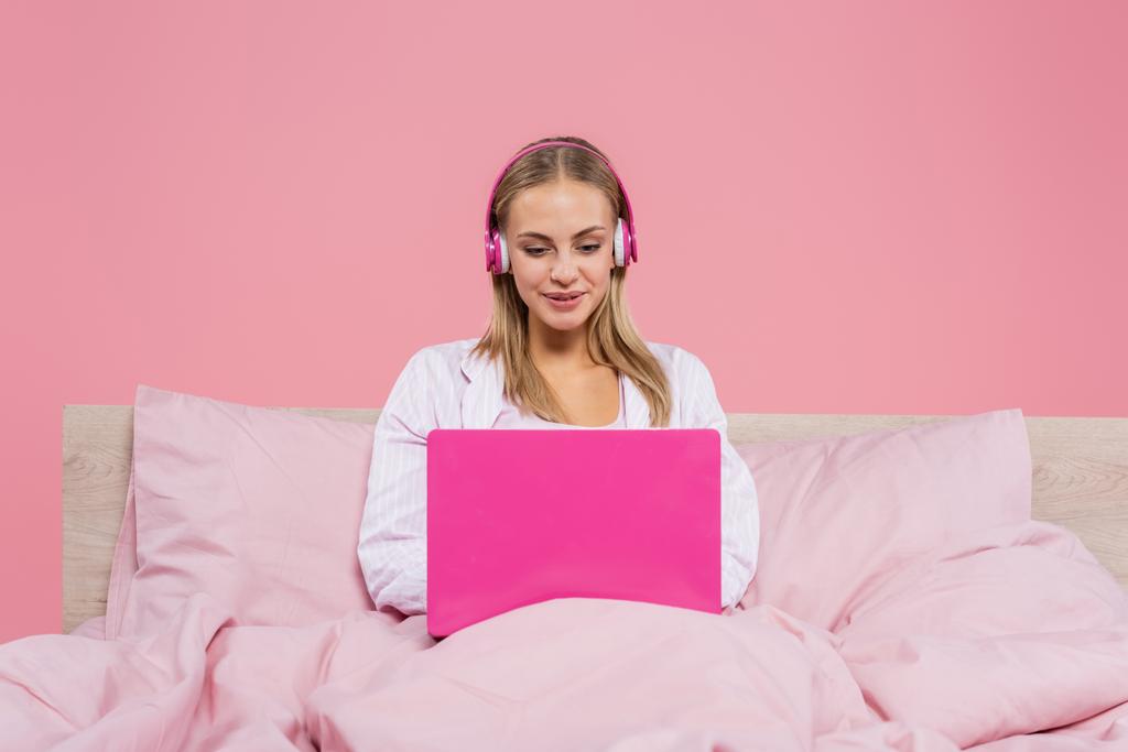 Freelancer sorridente em fones de ouvido usando laptop na cama isolado em rosa  - Foto, Imagem