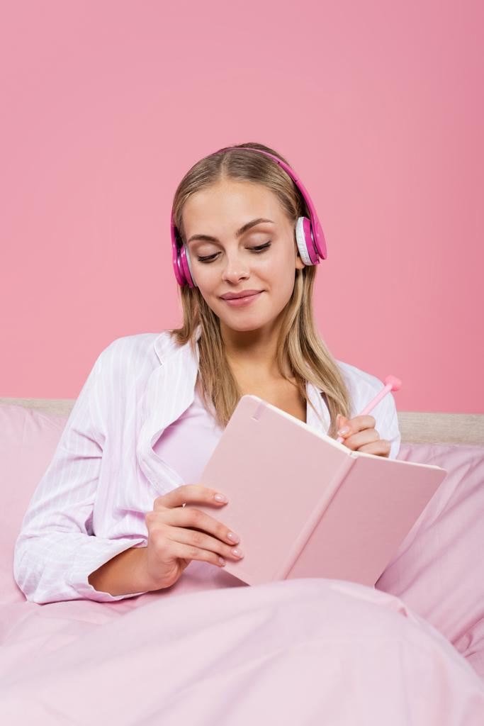 Χαμογελαστή ξανθιά γυναίκα με ακουστικά που γράφει στο σημειωματάριο στο κρεβάτι απομονωμένη σε ροζ  - Φωτογραφία, εικόνα