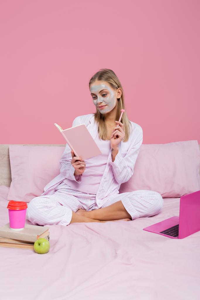 Blonde freelancer with face mask holding notebook near books, laptop and paper cup on bed isolated on pink  - Photo, Image