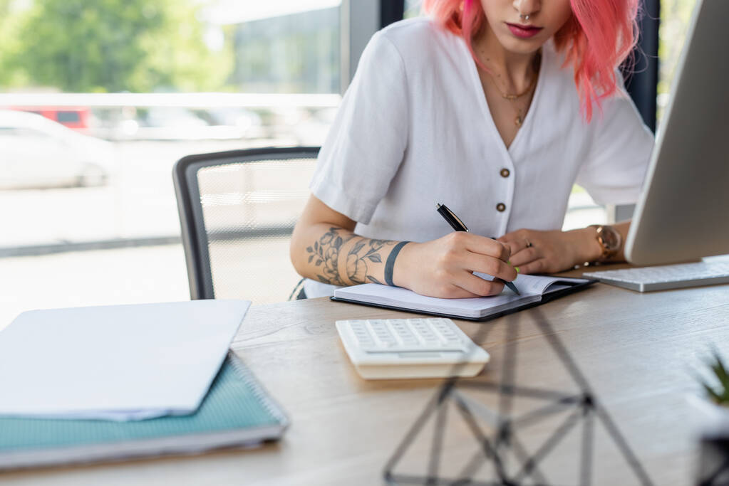 обрезанный вид проколотой деловой женщины с розовыми волосами, пишущей на блокноте в офисе - Фото, изображение