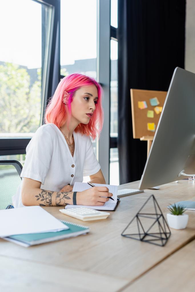 τρυπημένη επιχειρηματίας με ροζ μαλλιά γράφοντας στο σημειωματάριο, ενώ κοιτάζοντας οθόνη υπολογιστή  - Φωτογραφία, εικόνα