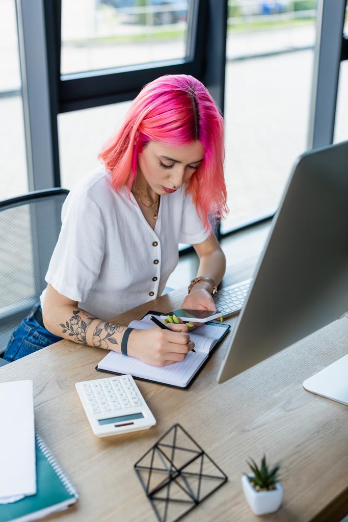 νέα επιχειρηματίας με ροζ μαλλιά γράφοντας στο σημειωματάριο, ενώ κρατώντας smartphone  - Φωτογραφία, εικόνα