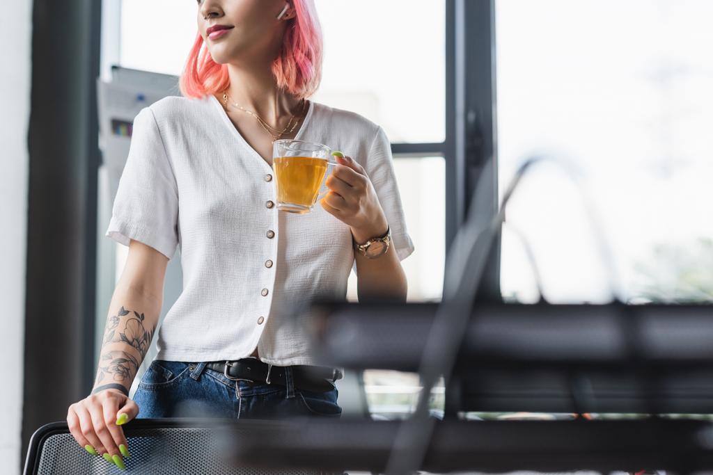 ostříhaný pohled na radostnou podnikatelku s růžovými vlasy a sluchátkem držící šálek čaje v kanceláři - Fotografie, Obrázek