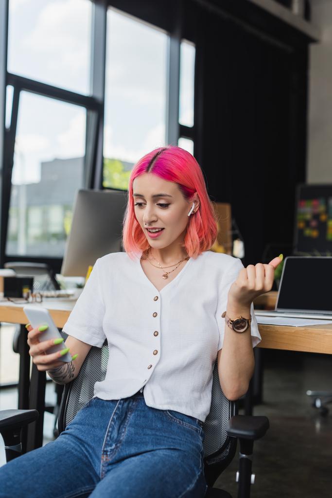 tetovaná mladá podnikatelka s růžovými vlasy a sluchátky pomocí smartphonu v kanceláři - Fotografie, Obrázek