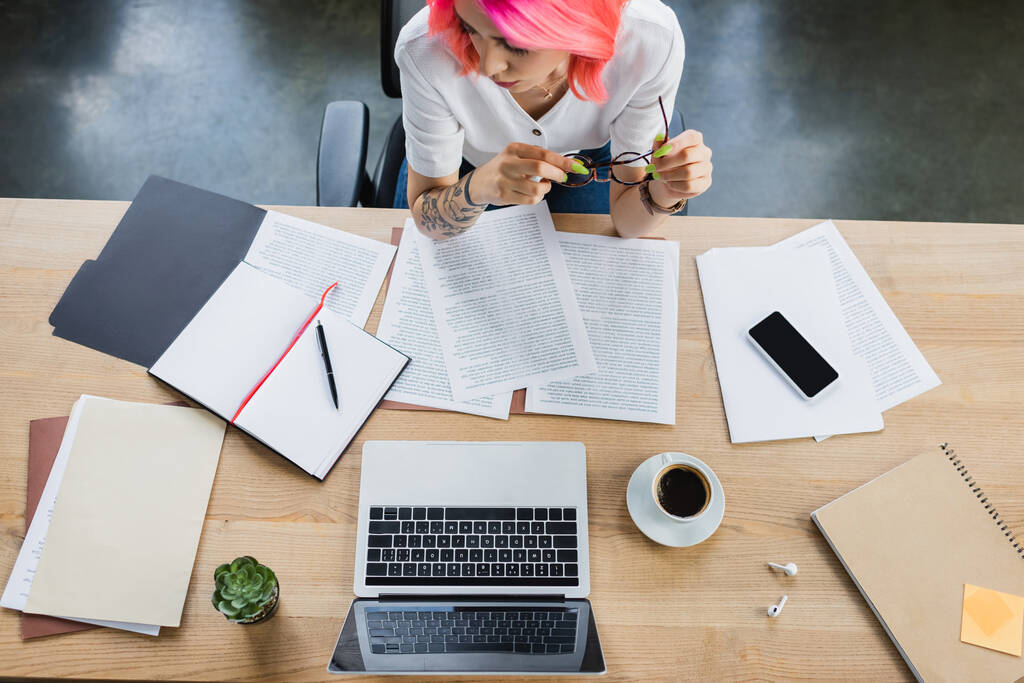 Draufsicht der Geschäftsfrau mit rosa Haaren, die eine Brille in der Nähe von Dokumenten und Gadgets auf dem Schreibtisch hält - Foto, Bild