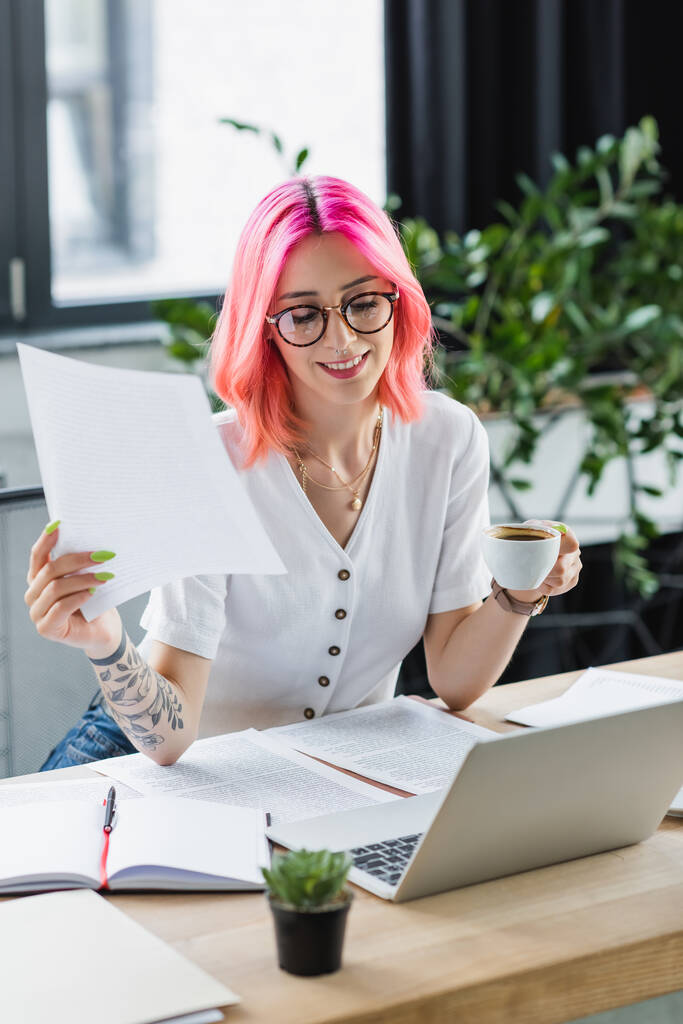 happy manager with pink hair holding cup of coffee near laptop and documents - Photo, Image
