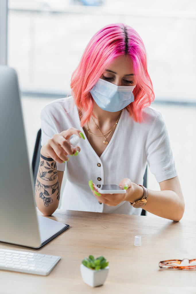 joven mujer de negocios en máscara médica rociando desinfectante en el teléfono inteligente en la oficina - Foto, Imagen